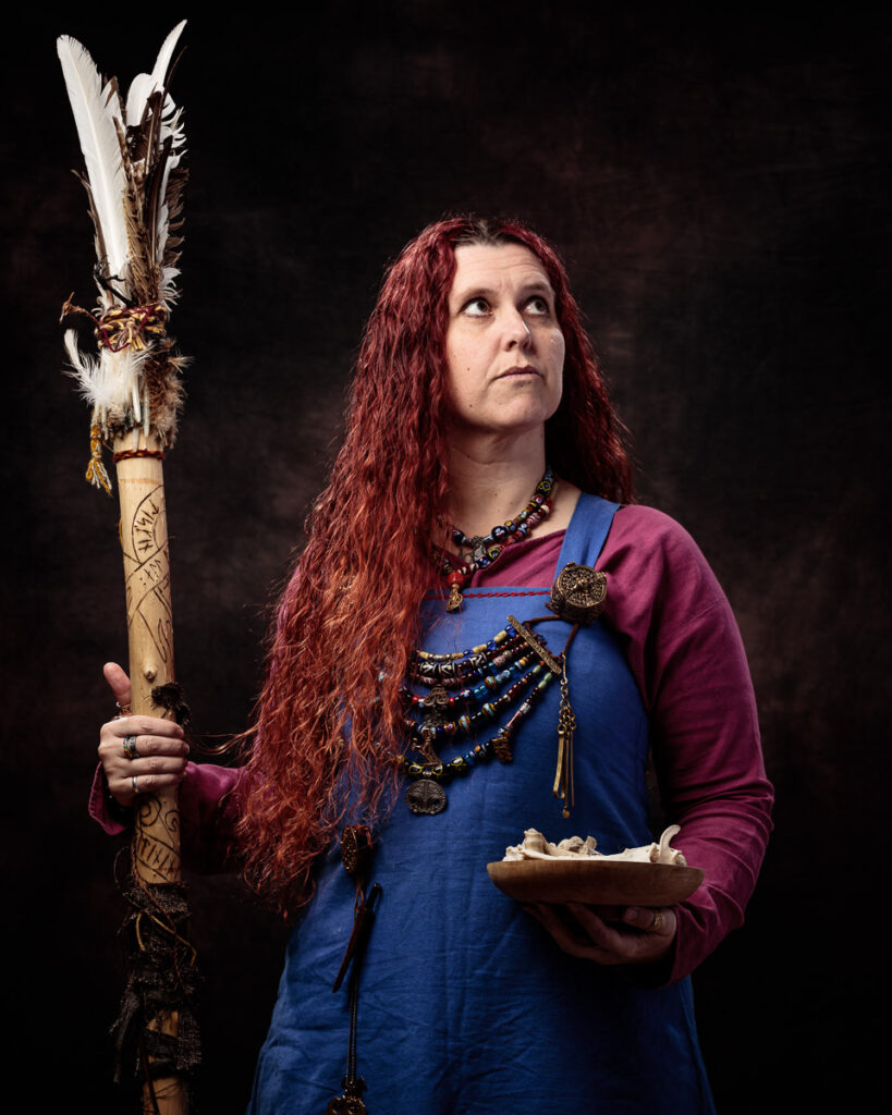 A person with long red hair wearing Viking-style clothing holds a feathered staff and a small wooden bowl against a dark background.