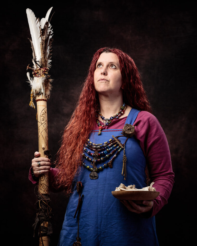 A person with long reddish hair wearing a blue tunic and burgundy overdress holds a wooden staff with white feathers and gazes upward against a dark background.