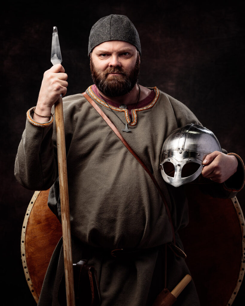 A bearded man in Viking costume holds an axe and helmet against a dark background.