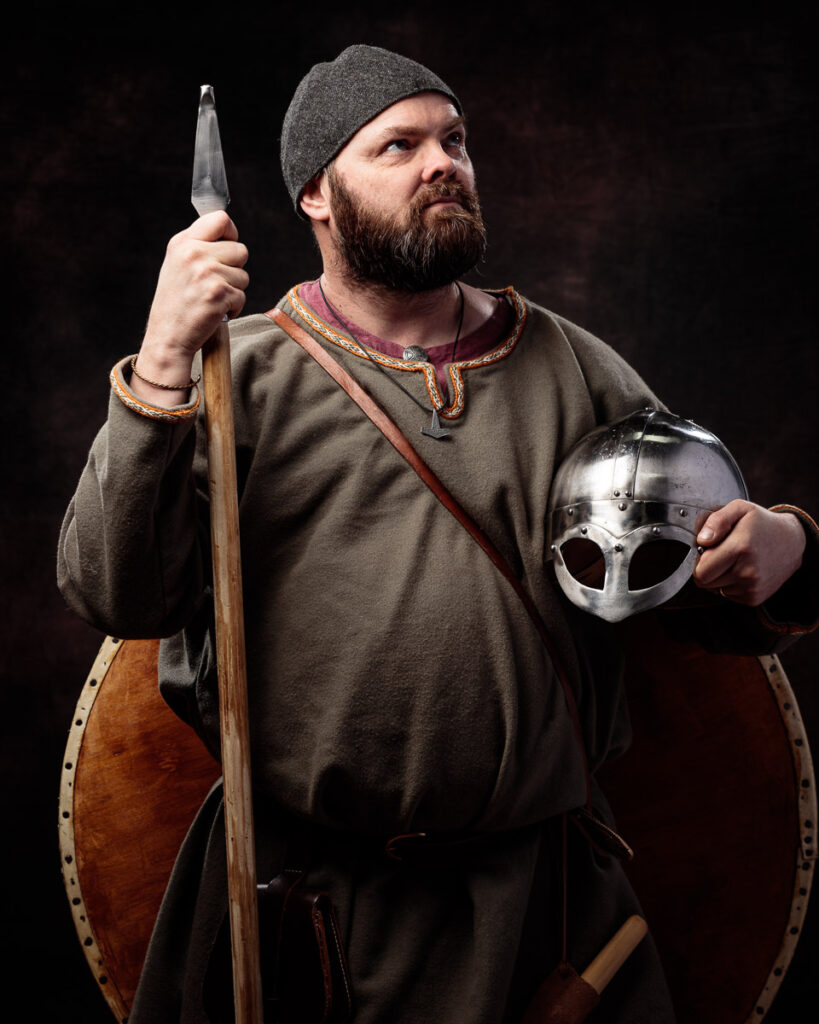 A bearded man in Viking-era costume holds an axe and shield against a black background, gazing upward pensively.