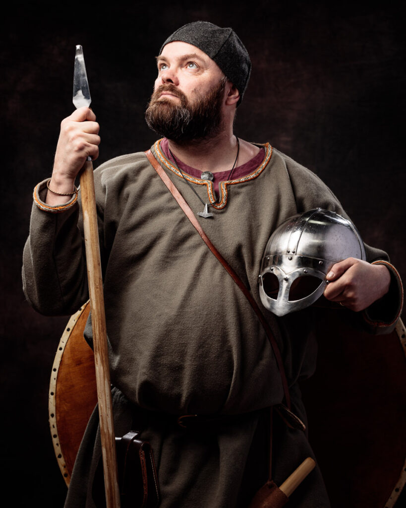 A bearded man in Viking-era clothing holds an axe and helmet against a dark background.