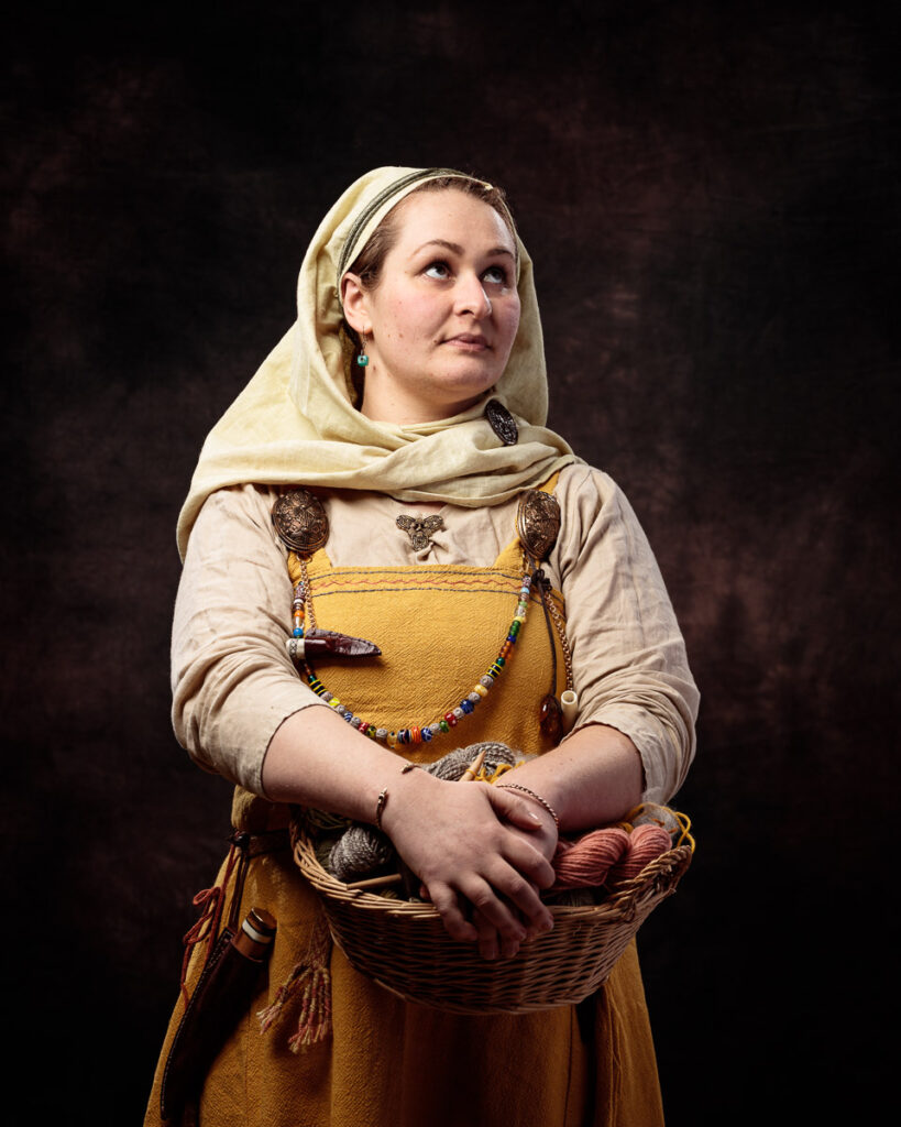 A woman wearing traditional Slavic folk costume with a mustard-yellow dress, cream linen shirt, and headscarf holds a wicker basket against a dark background.