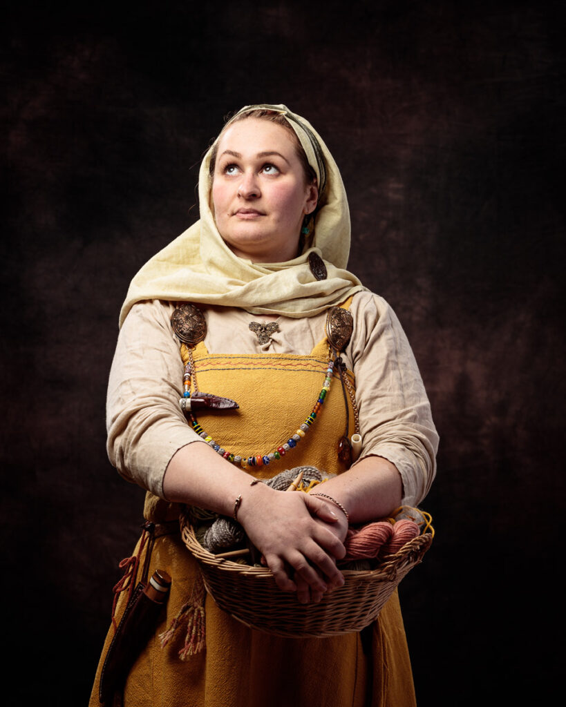 A woman in historical Norse or Viking-era clothing holds a wicker basket against a dark background, looking upward thoughtfully.