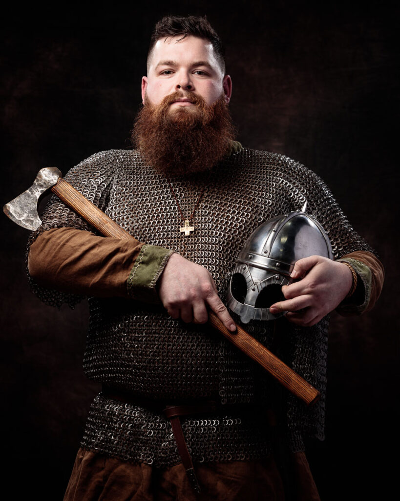 A bearded man in medieval chainmail holds an axe and helmet against a dark background.