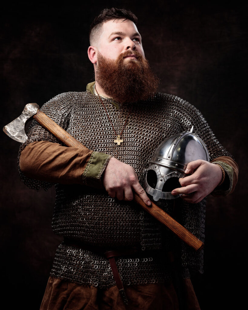 A bearded man in medieval warrior costume holds an axe and helmet against a dark background.