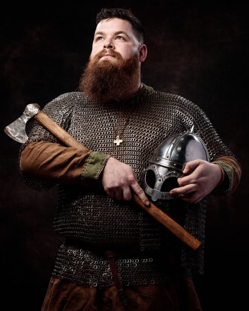 A bearded man in Viking-era armour holds an axe and helmet against a dark background.