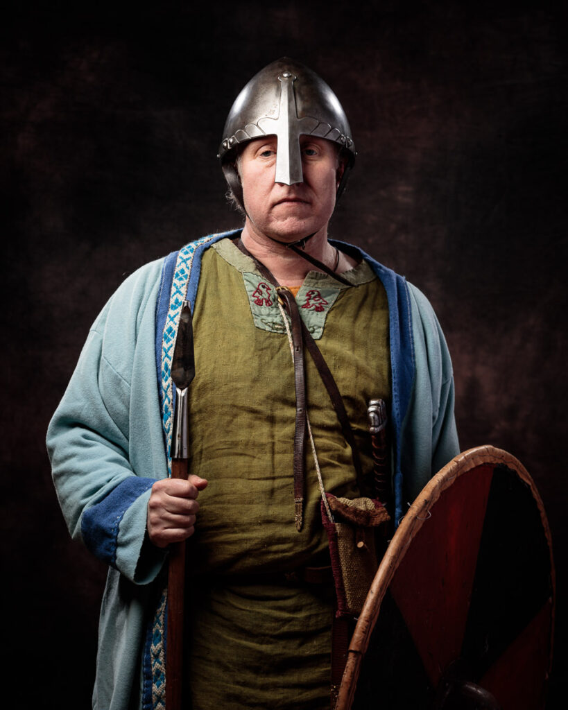 A man in Viking-era clothing and helmet holds a shield and axe against a dark background.