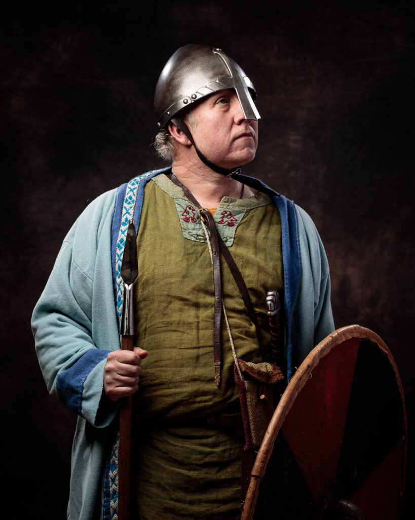 A man in Viking-era clothing and metal helmet holds a wooden shield against a dark background.