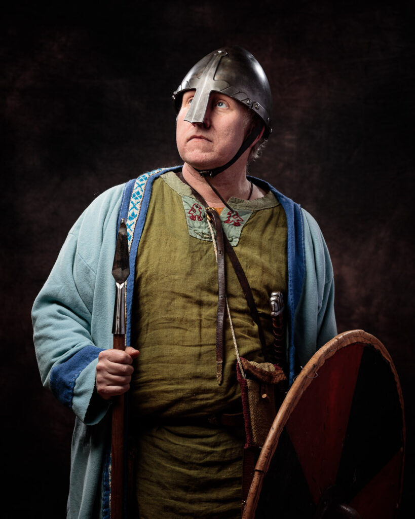 A man in Viking-era costume wearing a metal helmet and holding an axe, dressed in a tunic and cloak against a dark background.