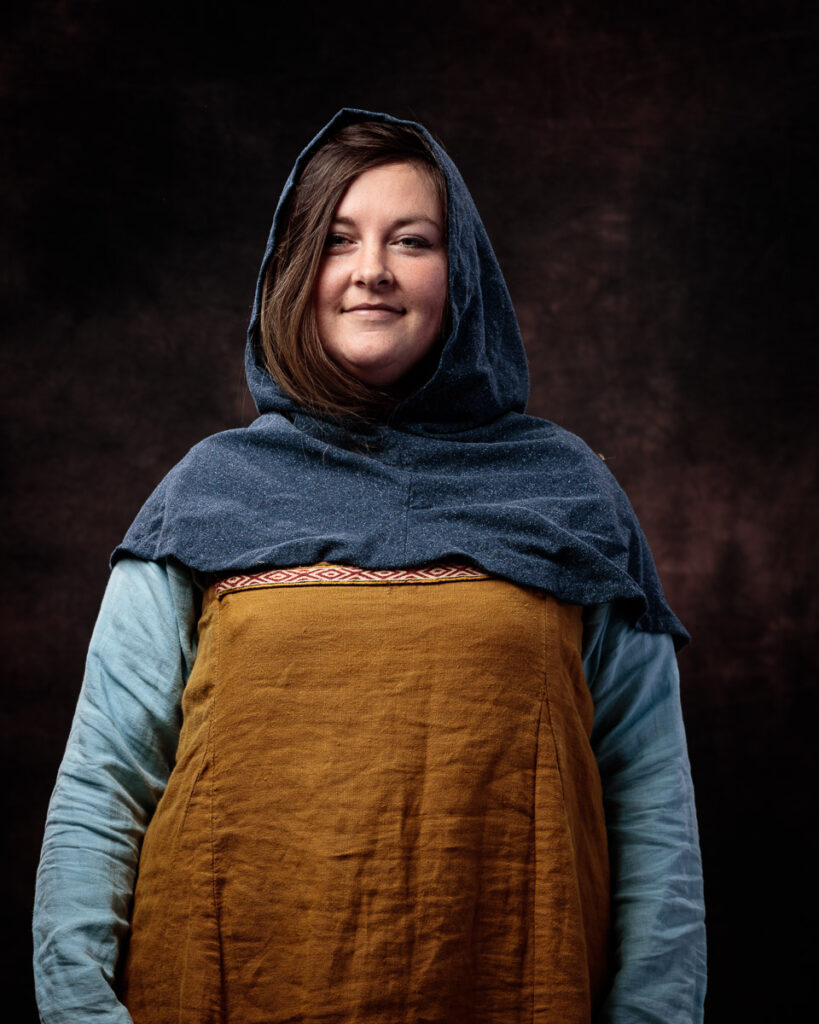 A woman wearing medieval-style clothing with a dark blue hood, light blue undershirt, and mustard-coloured tunic stands against a dark brown background.