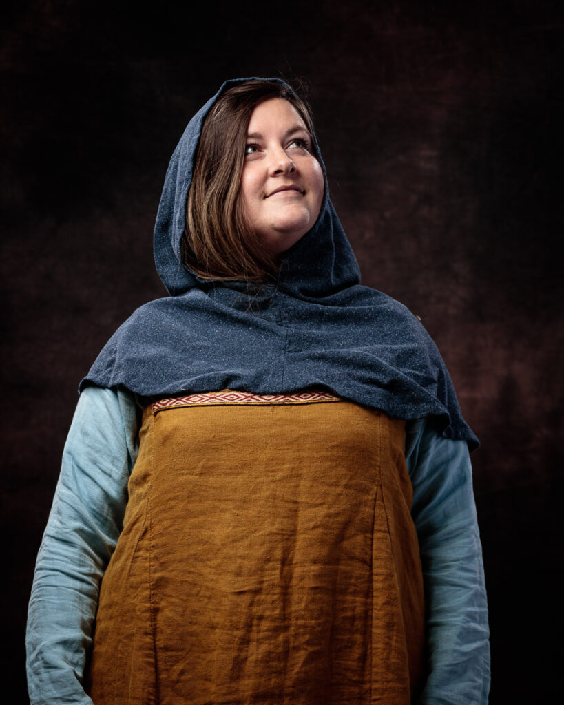 A young woman wearing historical Norse or Viking-inspired clothing stands against a dark background, looking upward thoughtfully.
