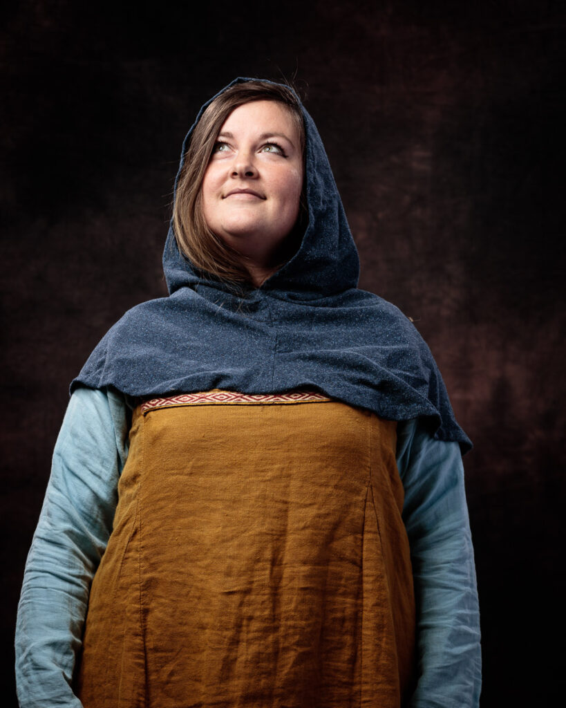 A woman wearing medieval-style clothing with a dark hood, tan tunic and blue undertunic, gazes upward against a dark background.