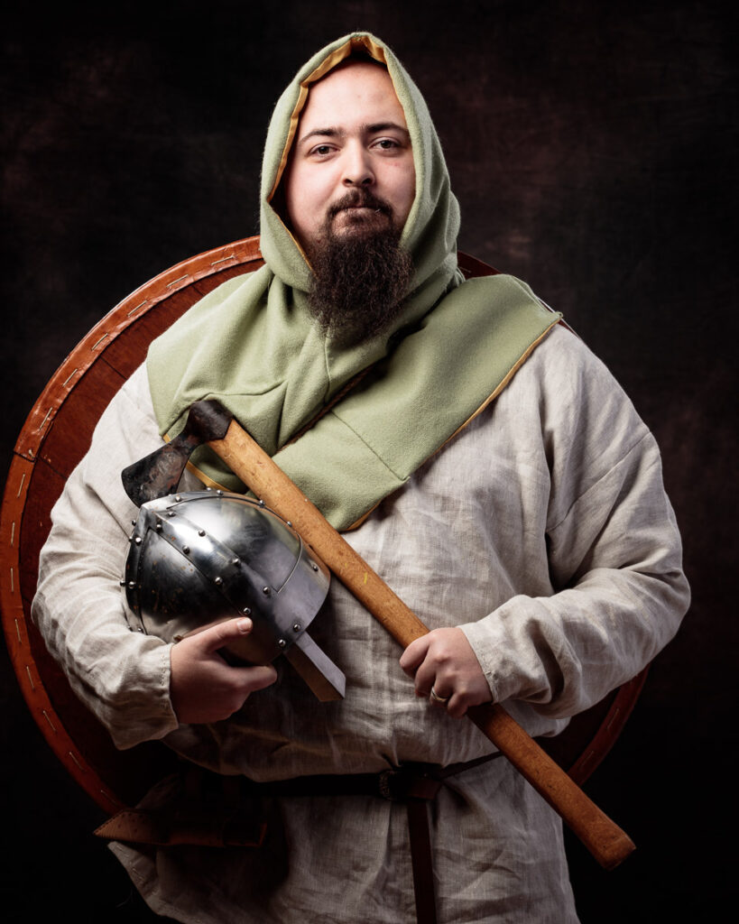 A bearded man in medieval clothing holds an axe and helmet against a dark background.