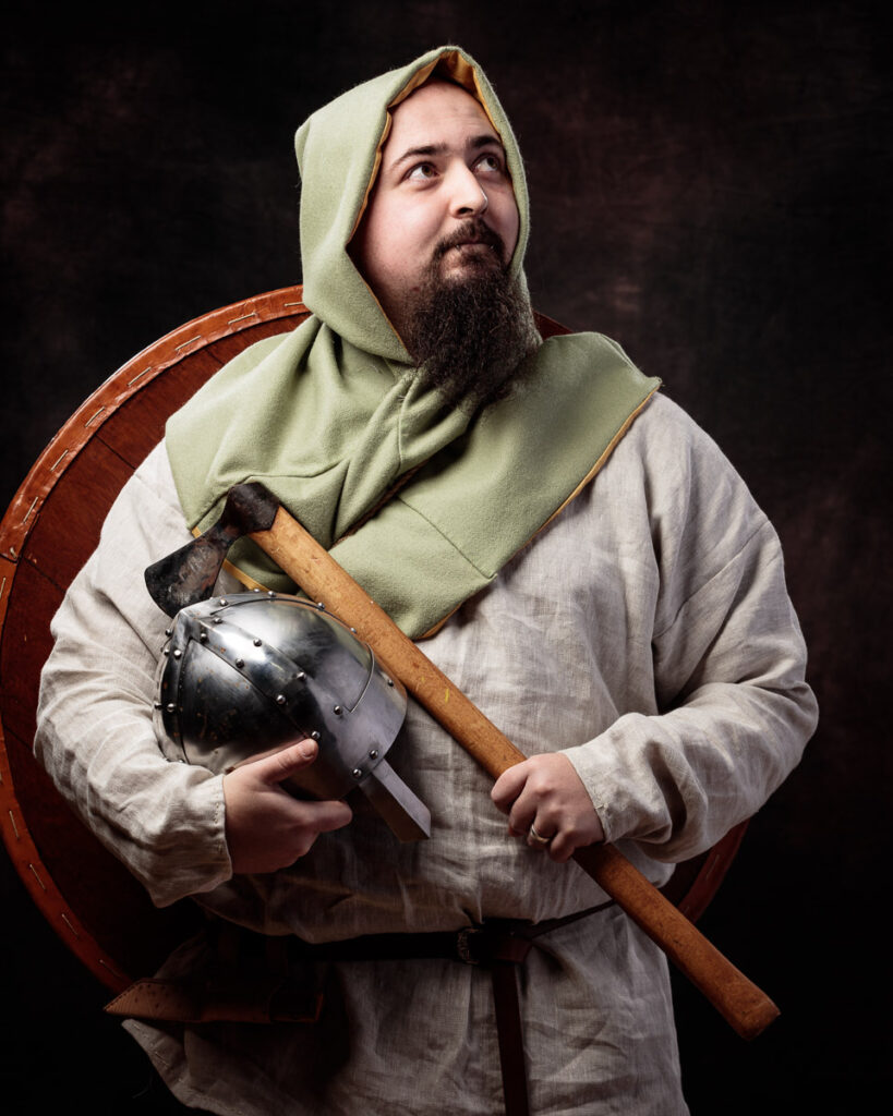 A bearded man in medieval costume holds an axe and helmet against a dark background, wearing a linen tunic and hood.