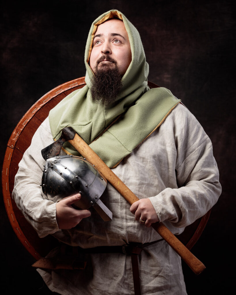 A bearded man in medieval clothing holds an axe and helmet against a dark background.