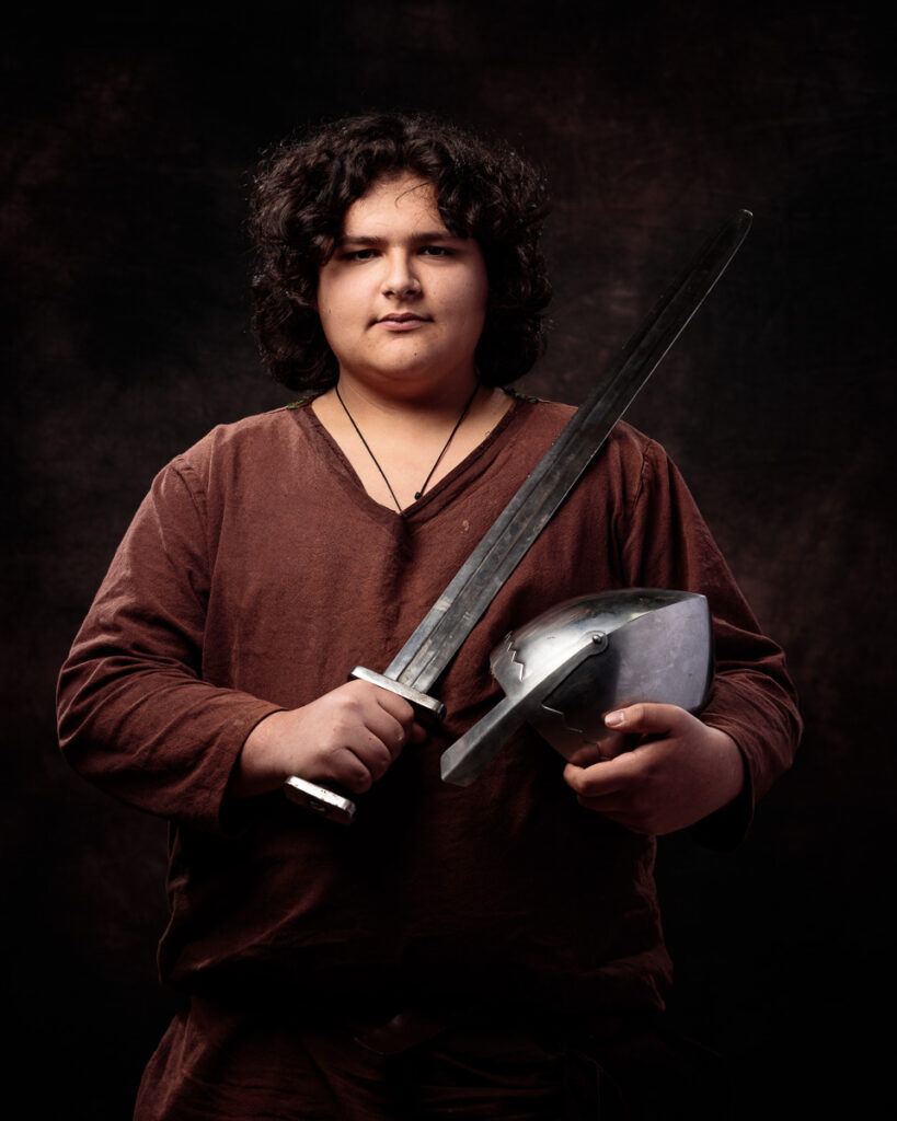 A young person with shoulder-length dark curly hair wearing a burgundy long-sleeved shirt holds a sword and shield against a dark background.