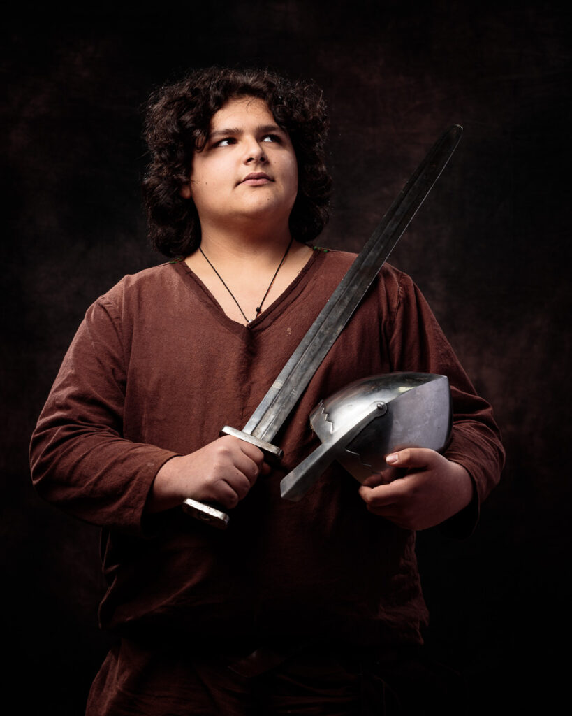 A young person with curly dark hair wearing a burgundy long-sleeved shirt holds a sword and shield against a dark background.
