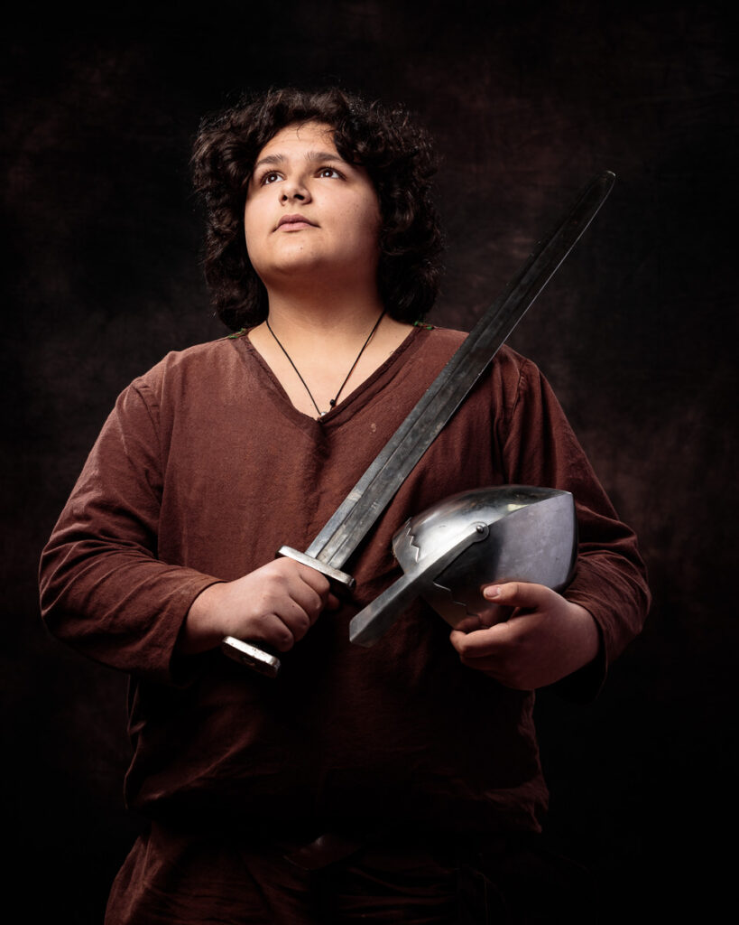 A young person with dark curly hair wearing a burgundy shirt holds a sword and shield against a dark background, looking upward.