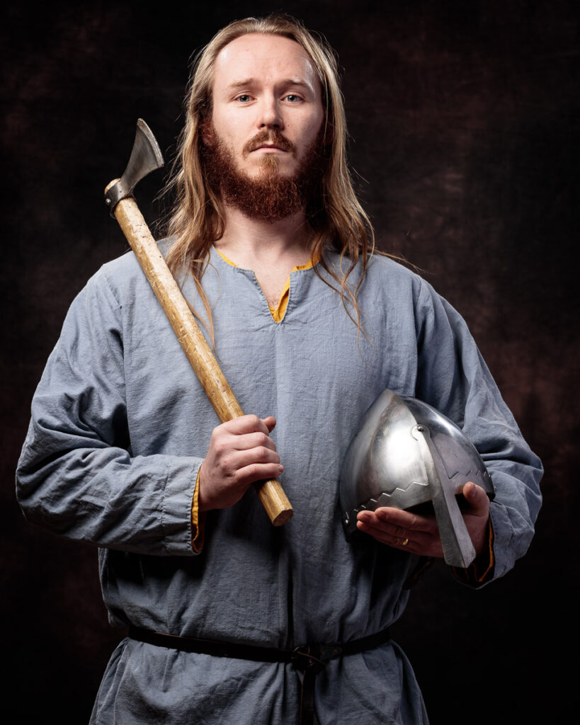 A man with long blonde hair and a full beard holds an axe and metal helmet against a dark background, wearing medieval clothing.