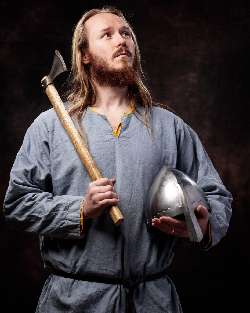 A bearded man with long hair wearing a grey tunic holds an axe and metal helmet against a dark background.