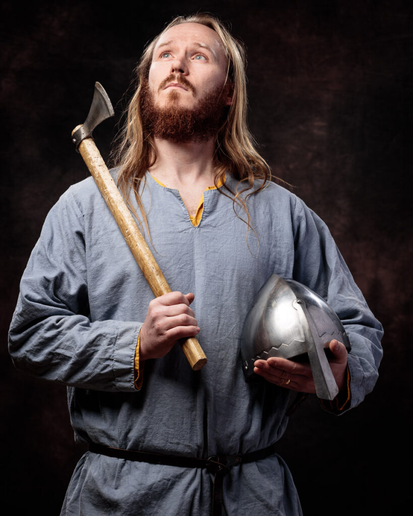 A man with long hair and a beard wearing a grey tunic holds an axe and metal helmet against a dark background.