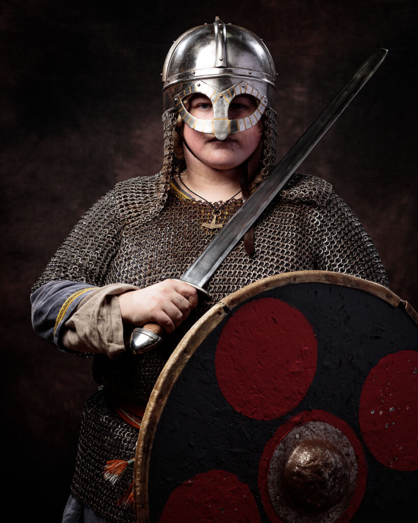A person wearing Viking-era armor and helmet holds an axe and shield against a dark background.