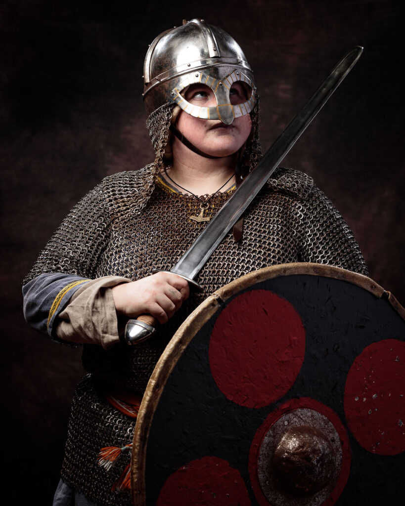 A Viking warrior in chainmail and helmet holds an axe and shield with red circular designs against a dark background.