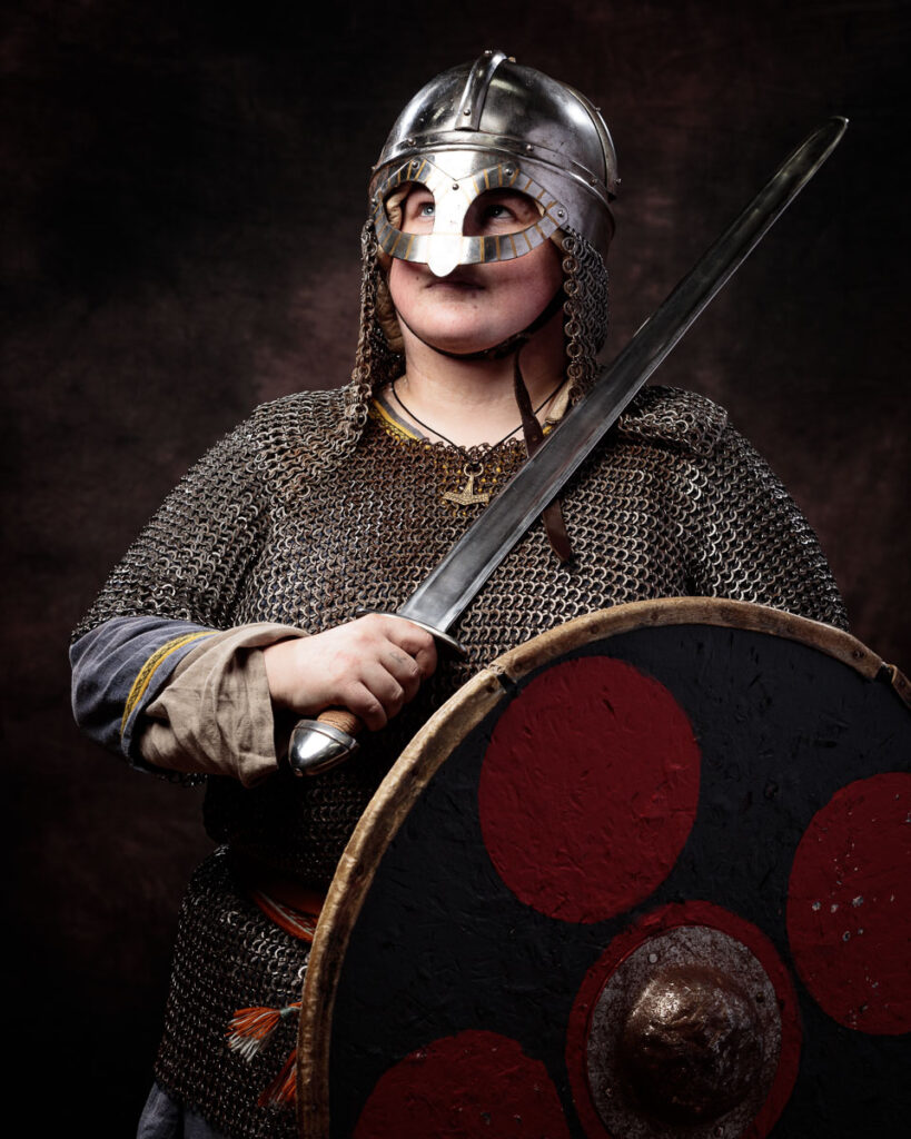 A person in Viking warrior costume wearing a polished metal helmet with face guard, chainmail tunic, and holding an axe and shield.