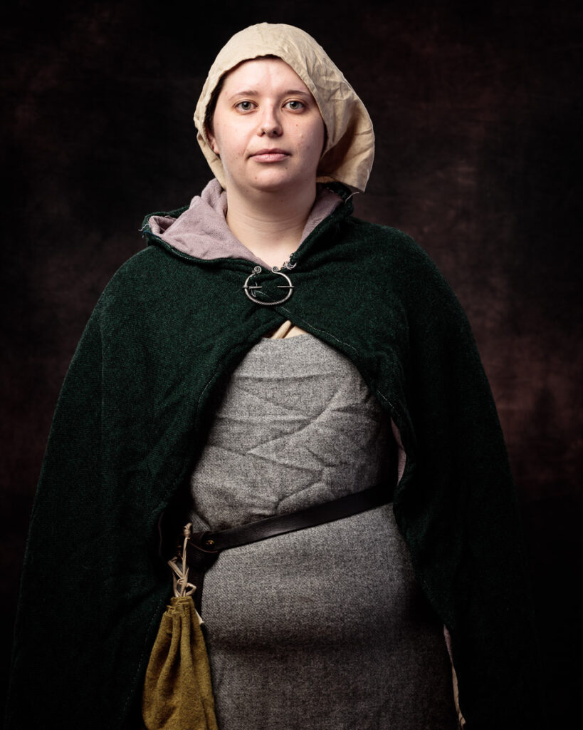 A woman in medieval-style clothing stands against a dark background, wearing a cream linen head covering, dark green cloak, and grey dress with a leather belt.