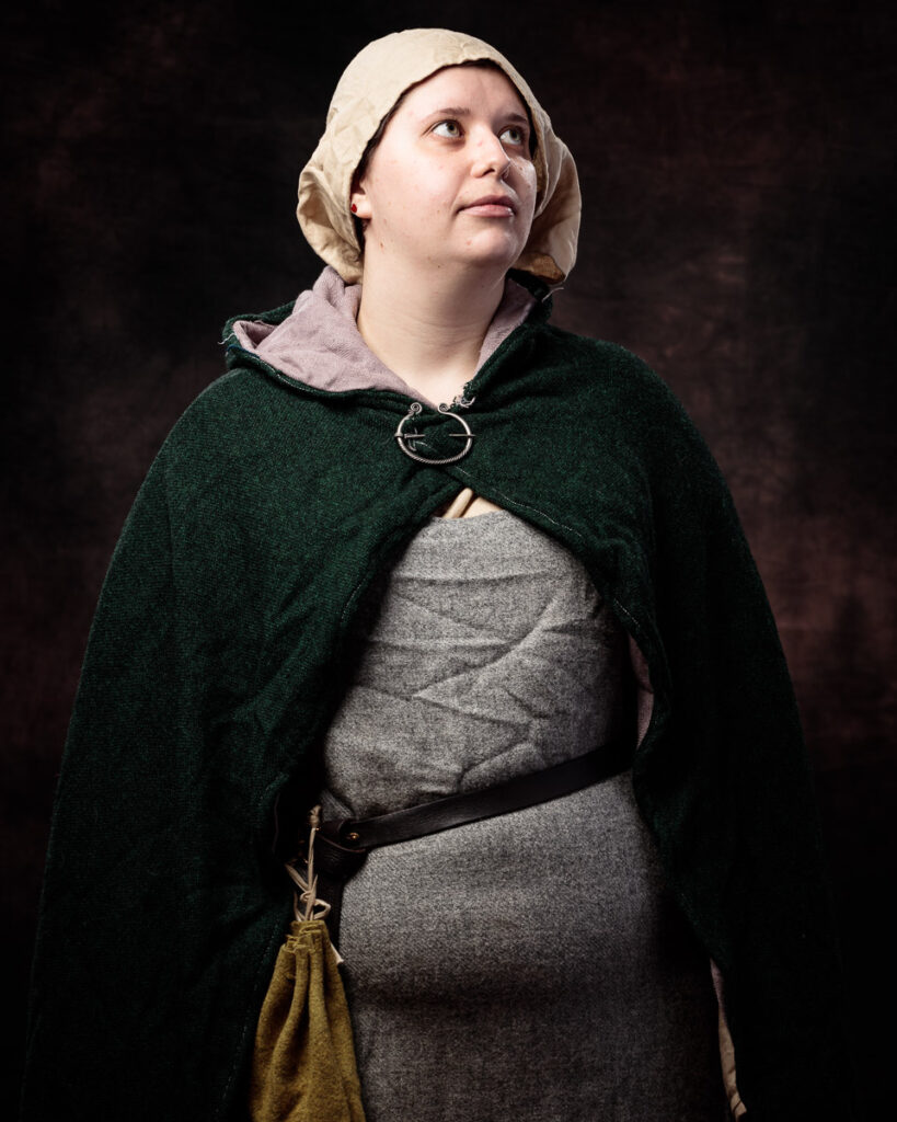 A person wearing medieval-style clothing looks upward against a dark background. They wear a cream linen coif, green wool cloak, grey tunic, and leather belt with a yellow pouch.