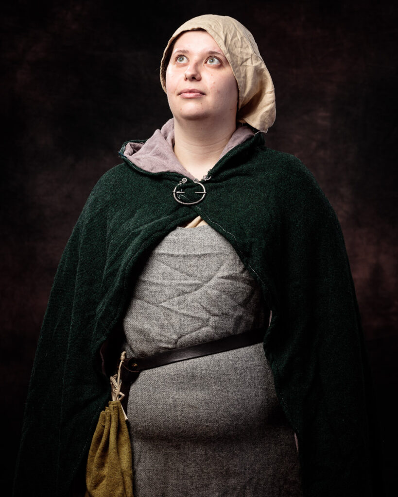 A woman in medieval-style clothing gazes upward. She wears a cream linen headdress, grey wool dress, dark green cloak, and holds a yellow pouch.