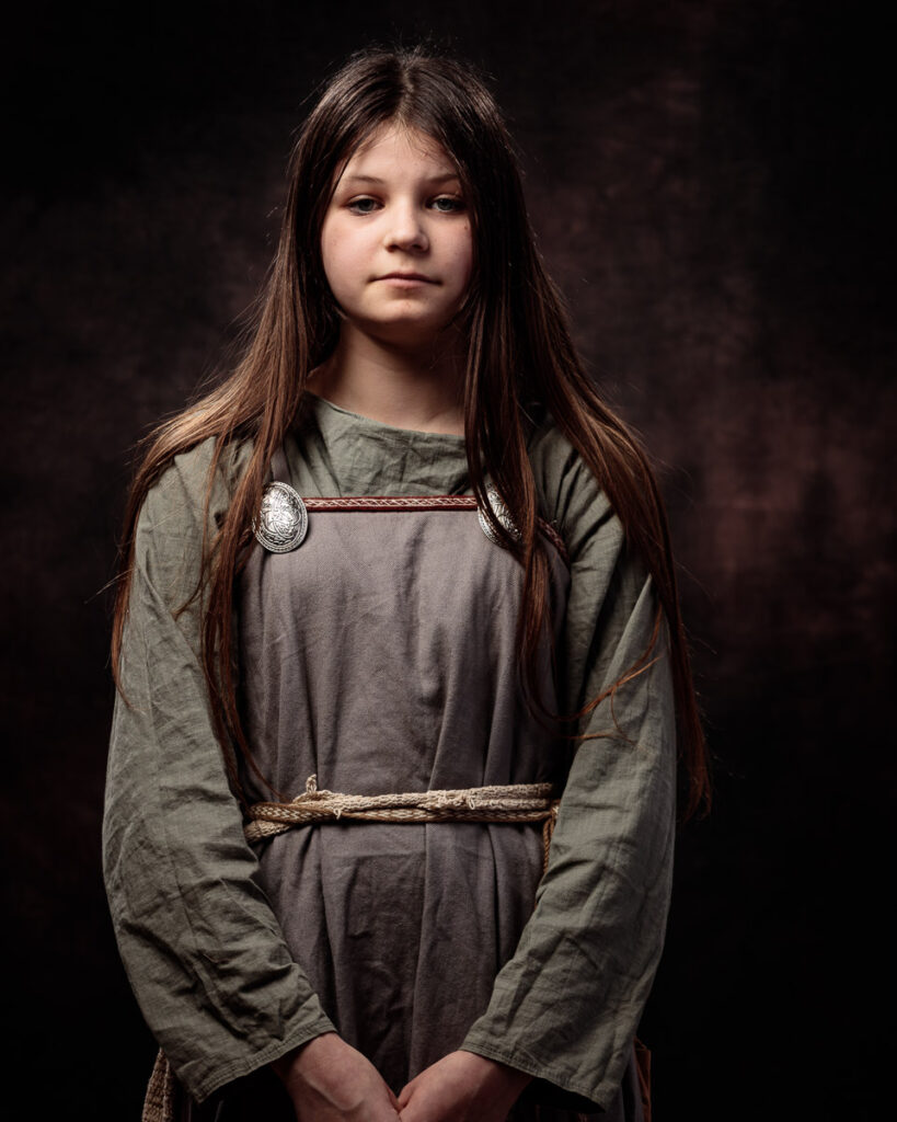 A young girl with long dark hair wearing a grey tunic with rope belts and decorative metal brooches, standing against a dark background.