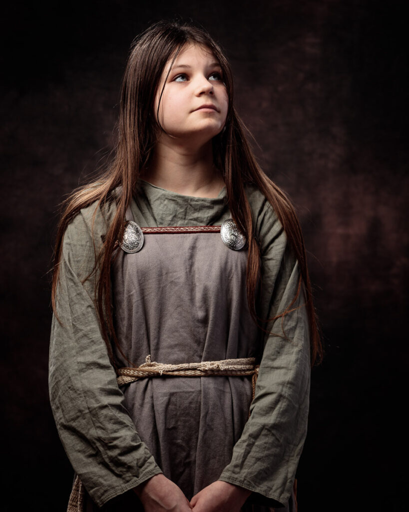 A young girl with long brown hair wears a grey Viking-style tunic with decorative brooches and rope belt against a dark background.