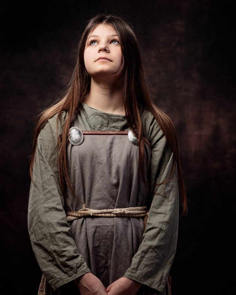 A young girl with long brown hair wearing a medieval-style tunic with decorative clasps, looking upward against a dark background.