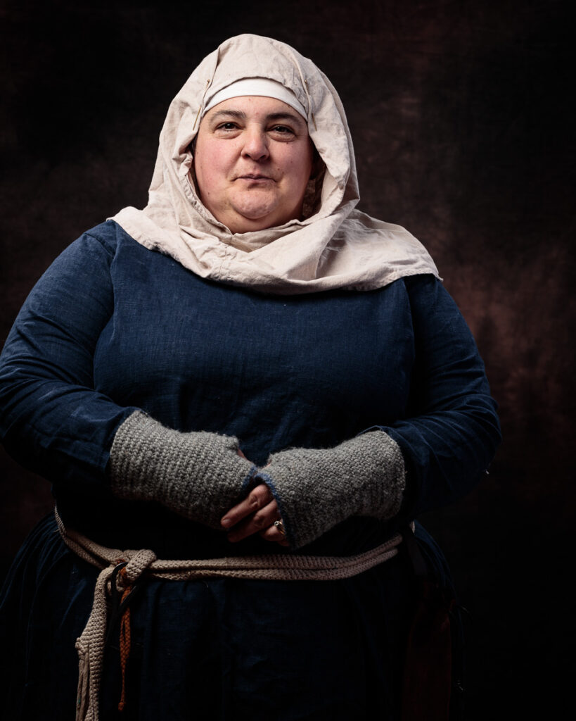 A woman wearing medieval-style clothing poses against a dark background. She wears a blue dress with a cream linen head covering and grey knitted mittens.