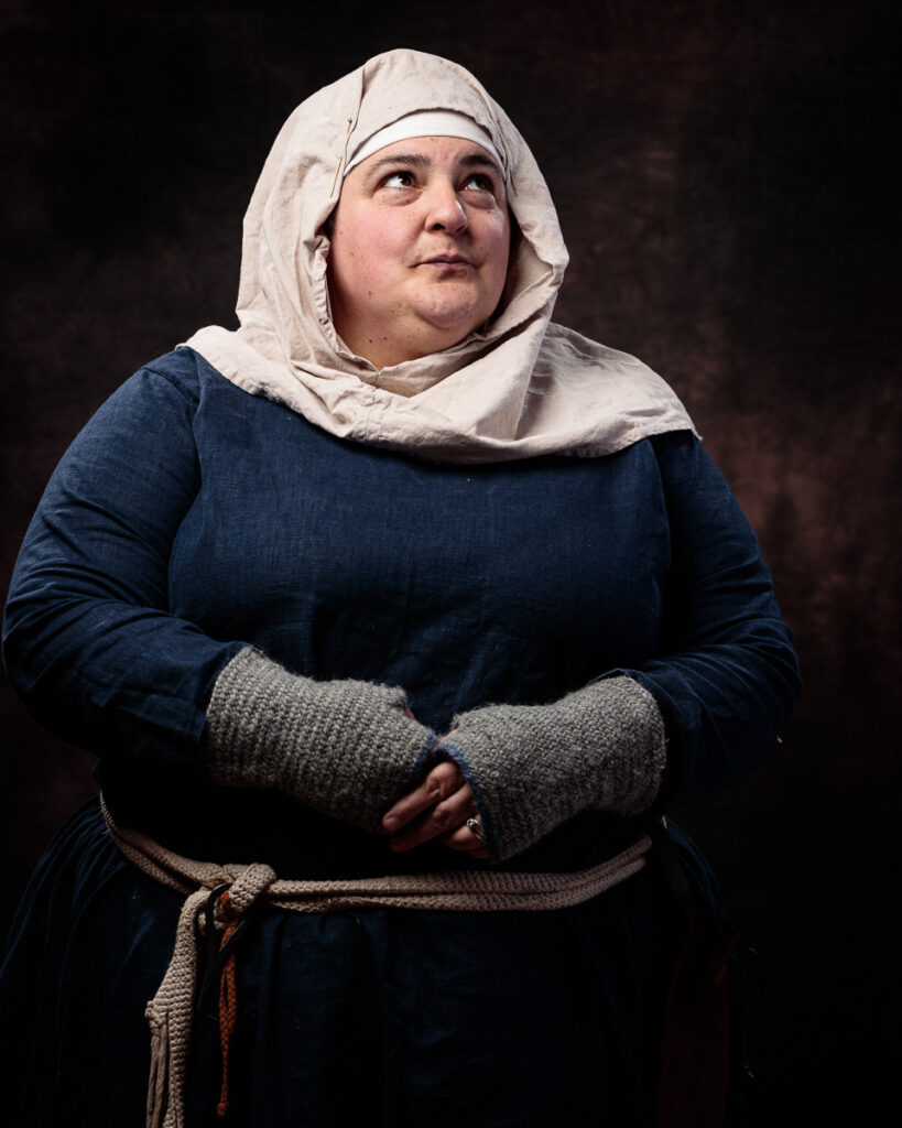 A woman wearing medieval-style clothing with a cream linen headscarf and dark blue dress looks upward against a dark background.