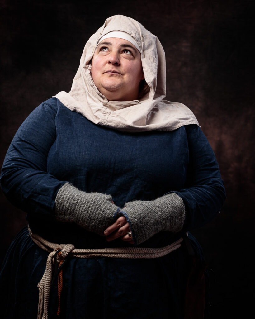 A woman in medieval clothing stands against a dark background, wearing a blue dress with a cream linen head covering and grey mittens.