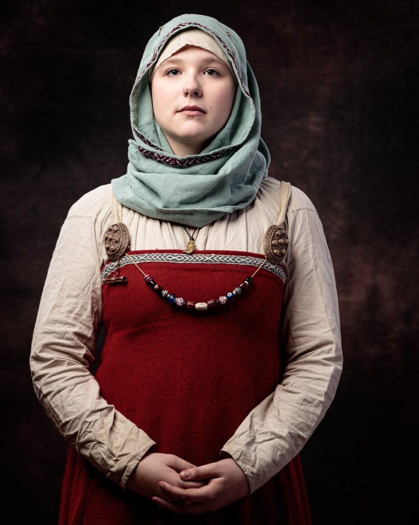A young woman wearing traditional Scandinavian folk costume with a red dress, linen underdress, and green headscarf stands against a dark background.