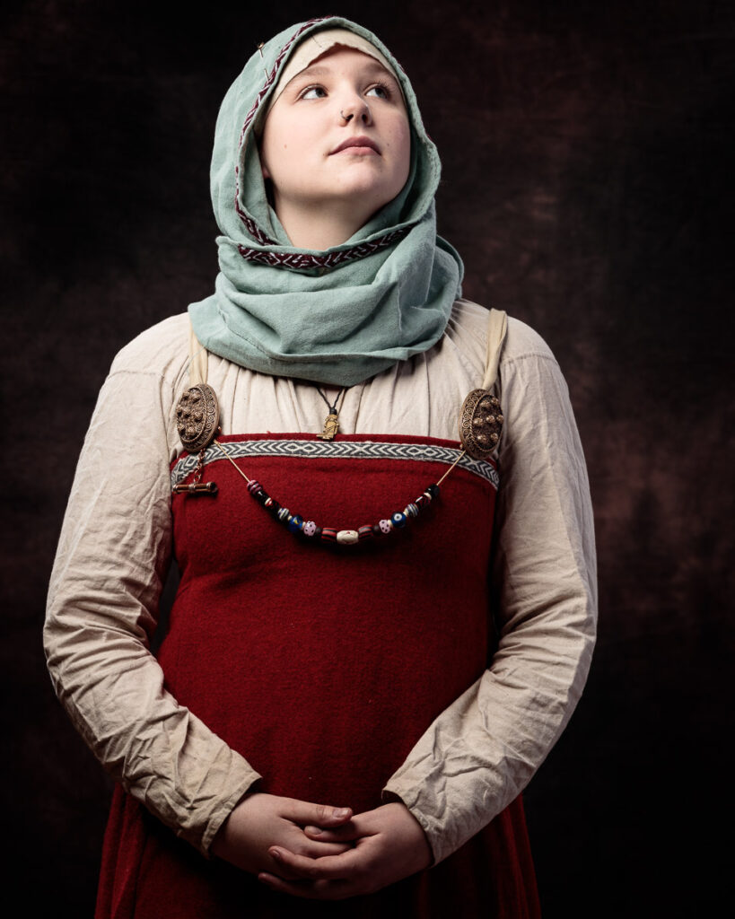 A young person wearing traditional Slavic clothing stands against a dark background, gazing upward. They wear a red sarafan dress with decorative beads, a cream linen shirt, and a pale green headscarf.