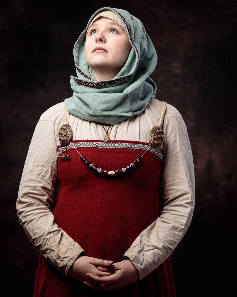 Young person wearing traditional Slavic folk costume with red dress, embroidered details, beaded necklace and grey-green headscarf, gazing upward against dark background.