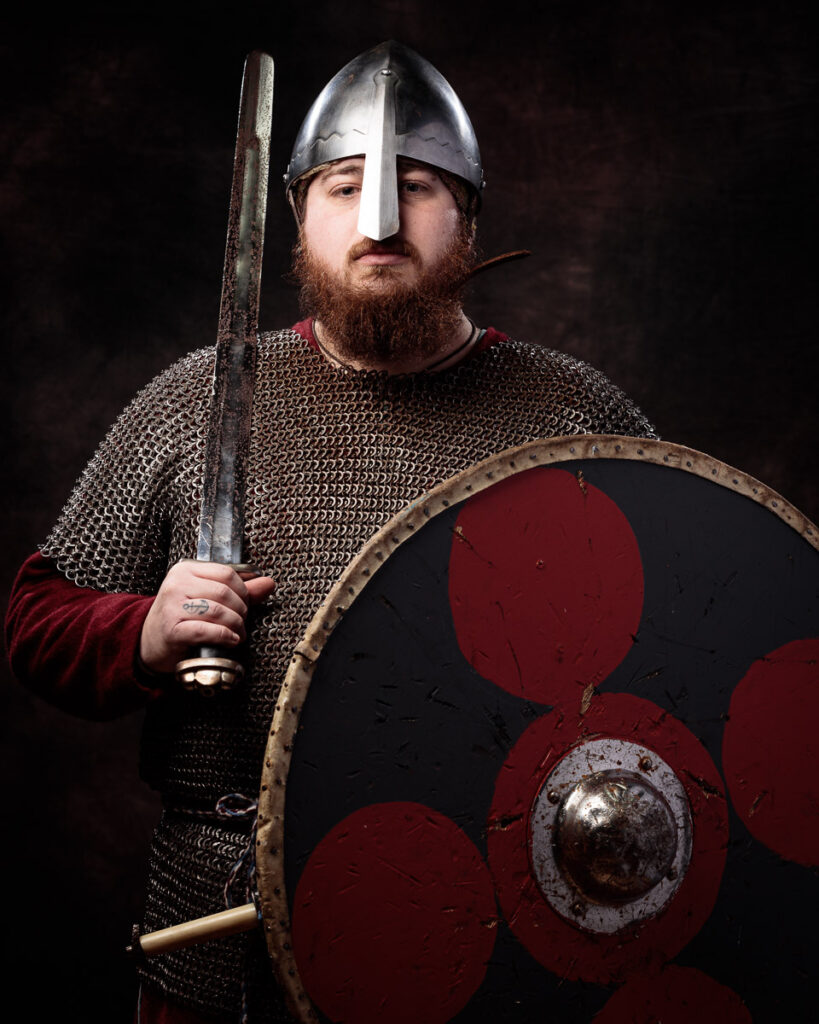 A bearded man in medieval warrior attire holds a sword and shield against a dark background.
