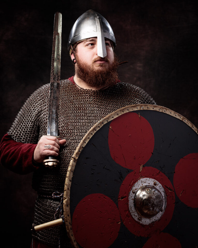 A bearded man dressed as a Viking warrior holds a sword and shield against a dark background.