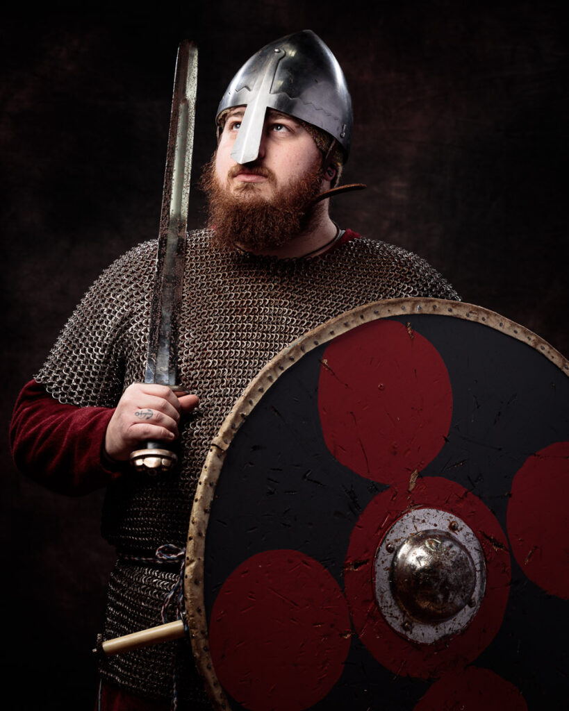 A bearded man in medieval warrior attire holds a sword and shield against a dark background.