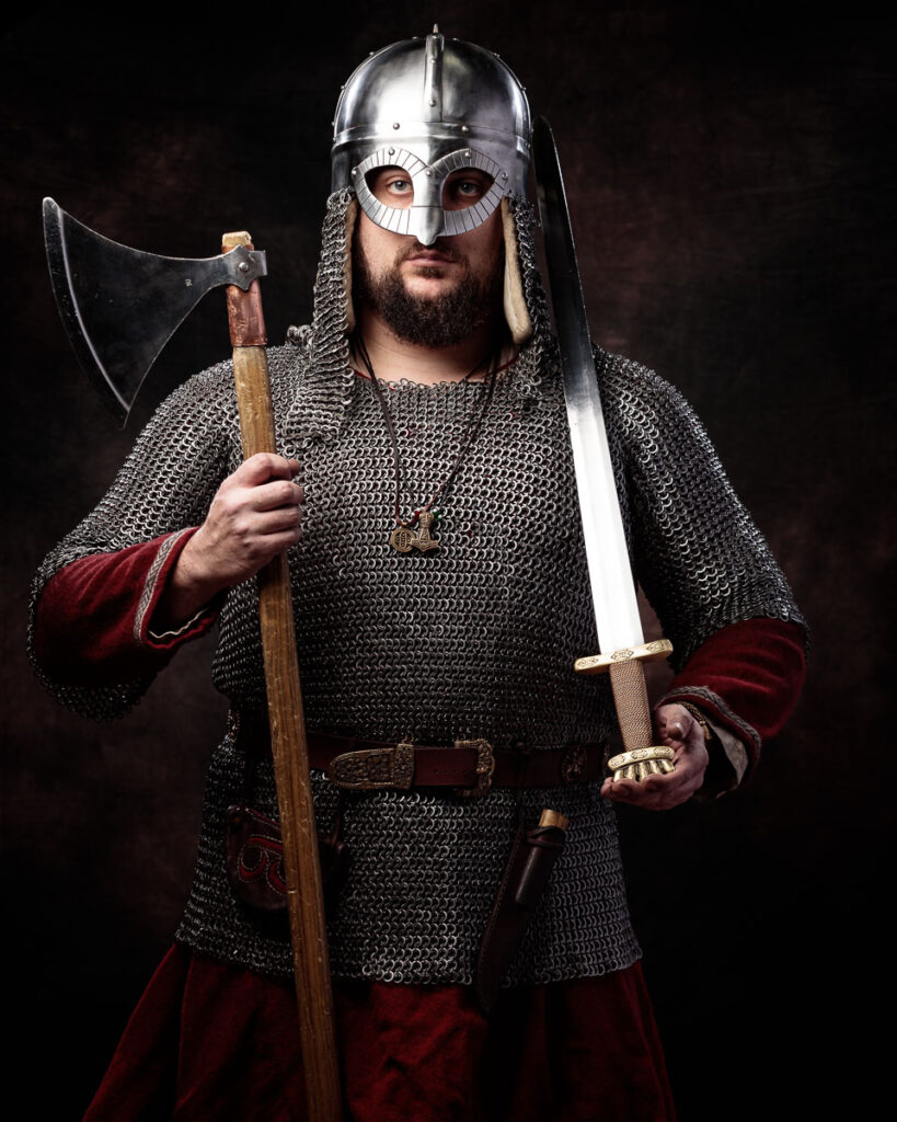 Viking warrior in chainmail and helmet holds an axe and sword against a dark background.
