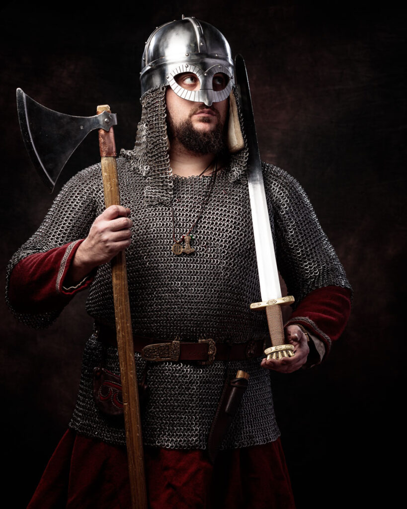 A bearded man wearing Viking-era armor and helmet holds an axe and sword against a dark background.