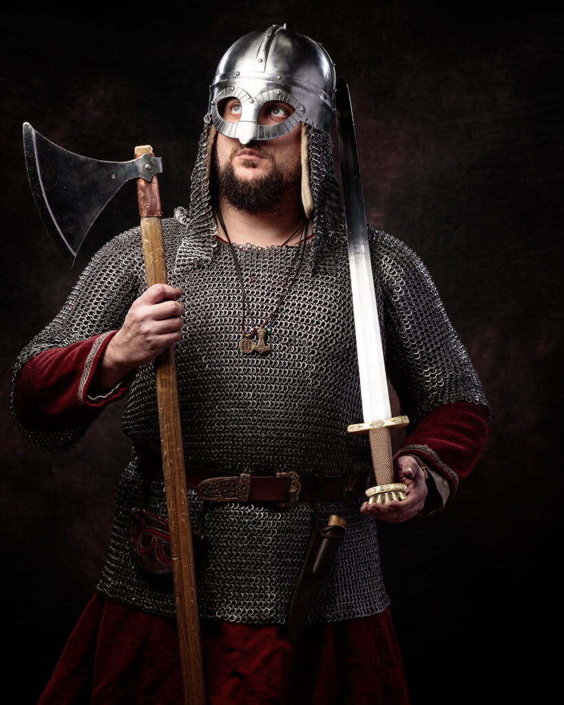 A bearded man in Viking warrior costume holding an axe and sword, wearing a metal helmet and chainmail shirt against a dark background.