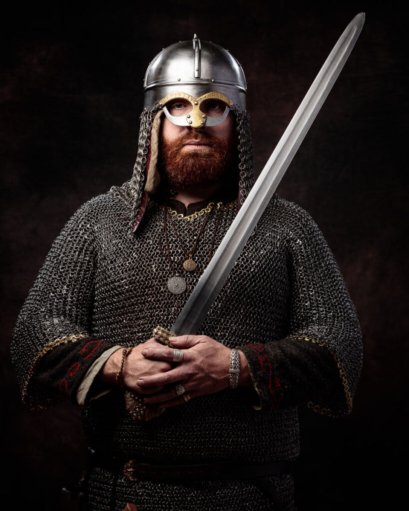 A bearded man in Viking warrior costume holds a sword, wearing a metal helmet with chainmail and protective armor against a dark background.