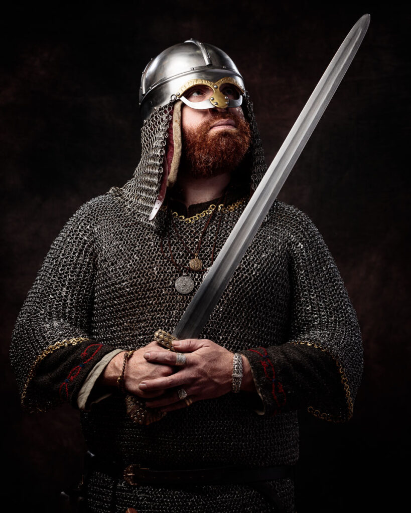 A bearded man in Viking warrior costume holds a sword, wearing a metal helmet, chainmail, and period jewelry against a dark background.