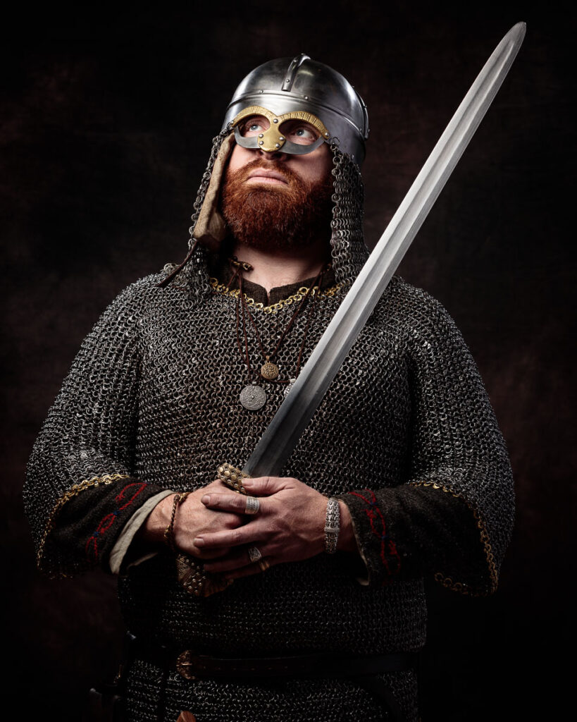 A bearded man in Viking warrior costume holds a sword, wearing a metal helmet with chain mail and ornate details against a dark background.