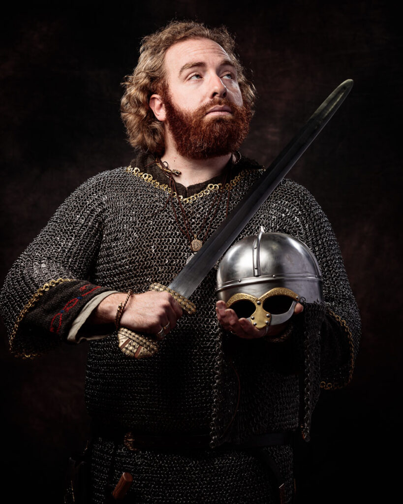 A bearded man in chainmail armour holds a sword and helmet against a dark background.